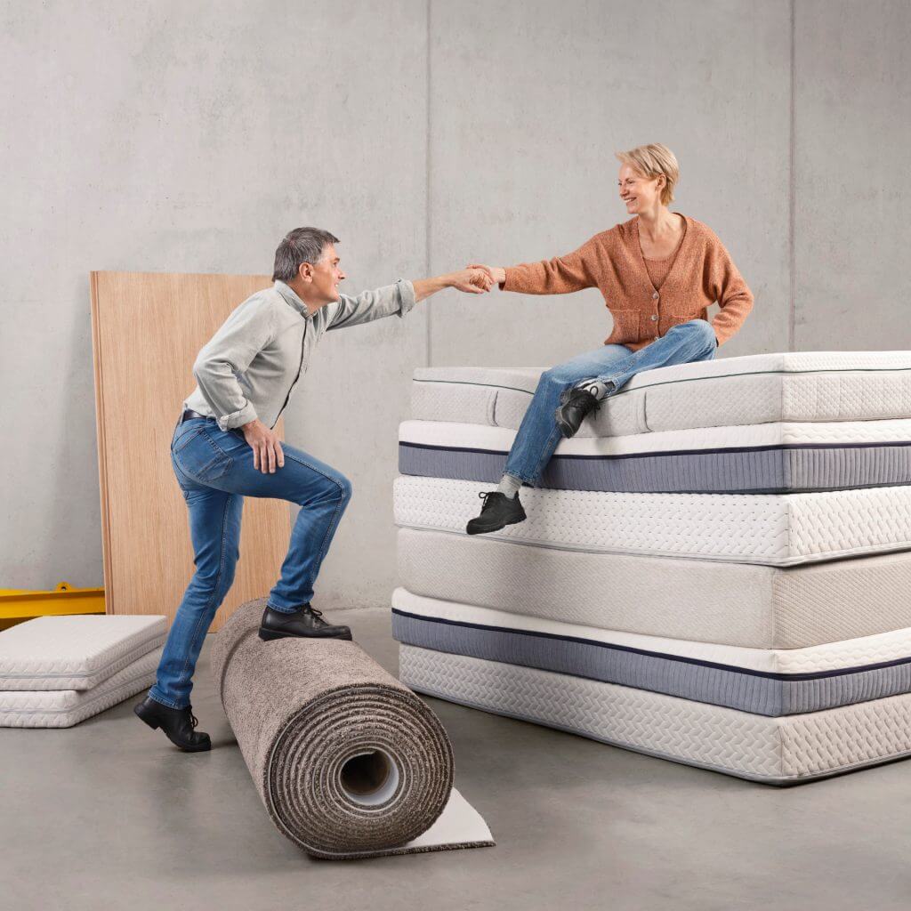 Man standing on mono-polyester carpet role with this hand reaching out and upwards to a woman sitting on a pile of mattresses designed for circularity. She stretches out her arm to the man to help him up.